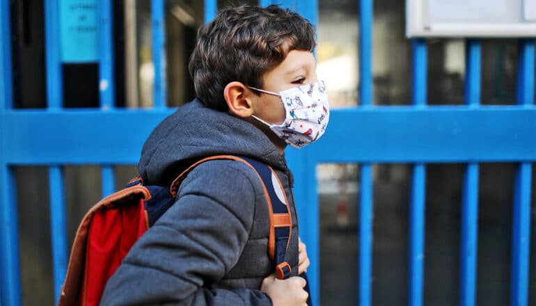 A young boy wearing a face mask walks to school with his backpack on.