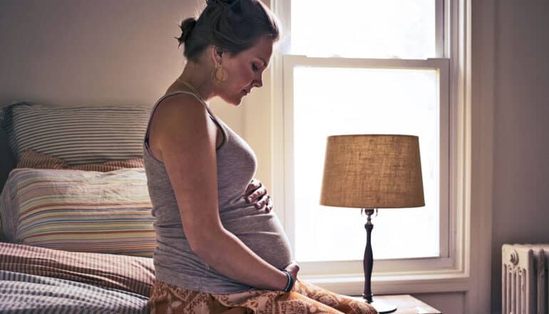 A pregnant woman sits on a bed looking concerned.