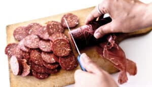 A person uses a knife to cut a sausage on a cutting board.