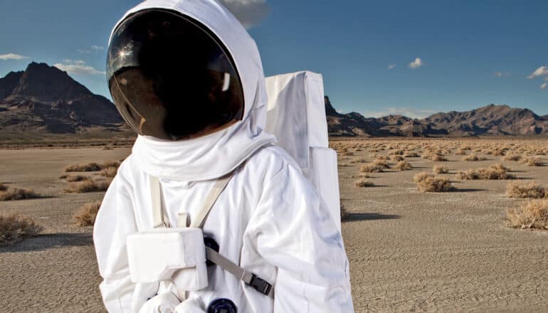 A person in an astronaut costume walks through a desert with mountains in the background.