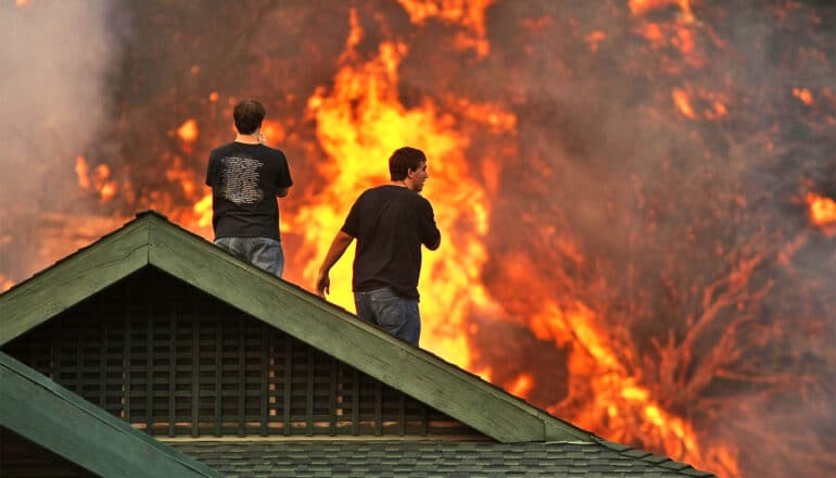 Two people stand on the roof of a house watching a nearby wildfire burn.