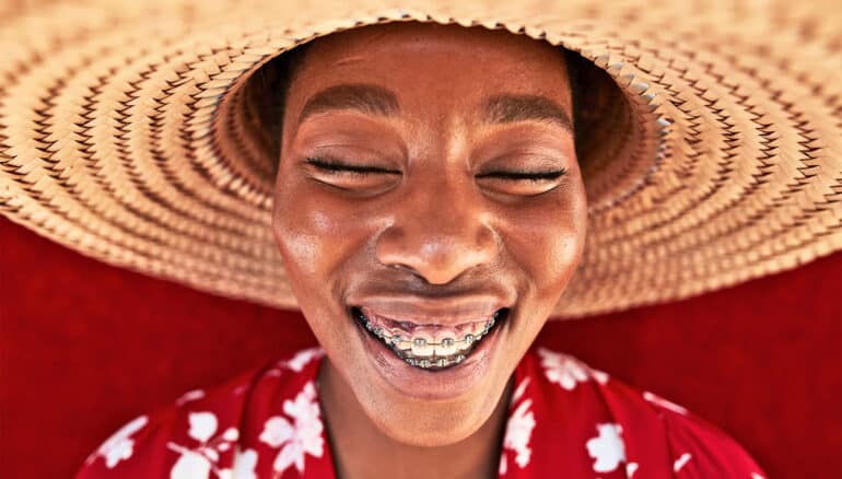 A person wearing braces and a large hat smiles widely.