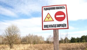 A sign warns of radiation with a brown field and trees in the background where a village once was.