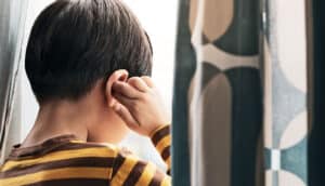 A young boy touches his ear while looking out a window.