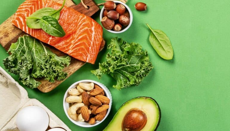 Foods that make up a keto diet, including avocado, salmon, nuts, eggs, and greens, on a green background.