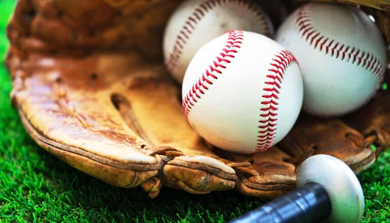 A baseball glove with three baseballs in it sitting on a field next to a bat.