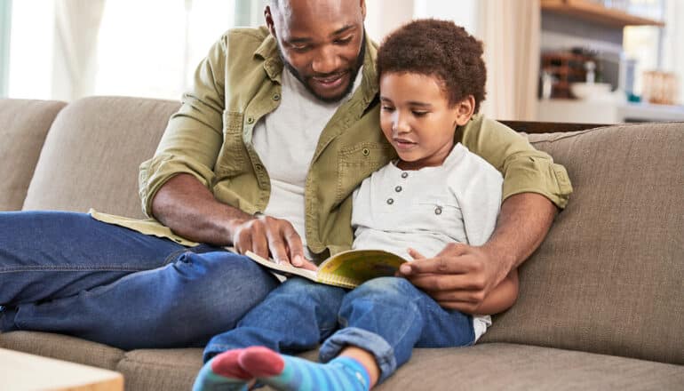 A father reads a book with his son while sitting on a couch.