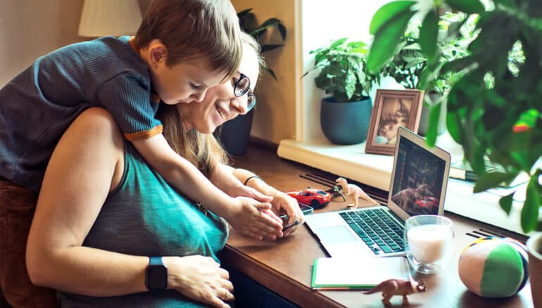 A pregnant woman sits at a desk in front of a computer as her son hugs her from behind.