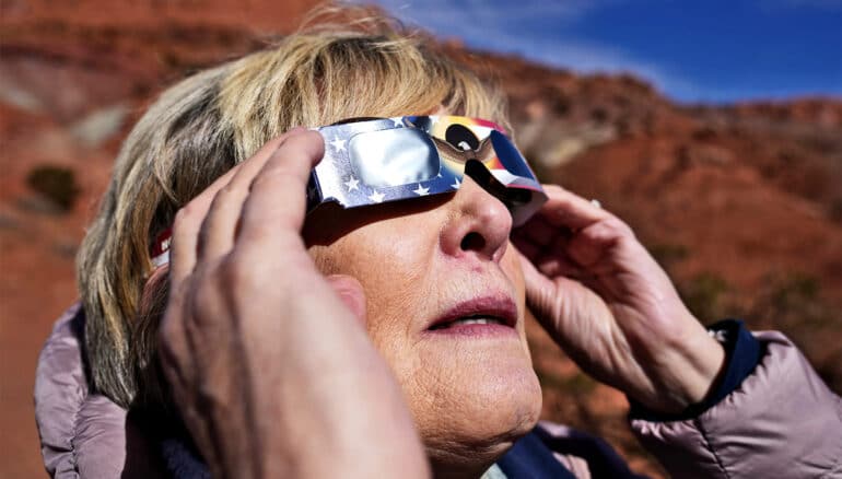 A woman holds solar eclipse glasses to her face as she looks up to the sky.