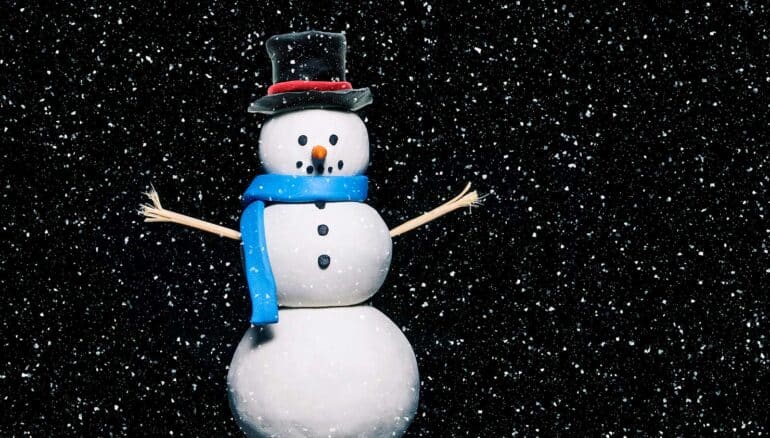 A toy snowman sits against a black background with falling snowflakes that look like stars.