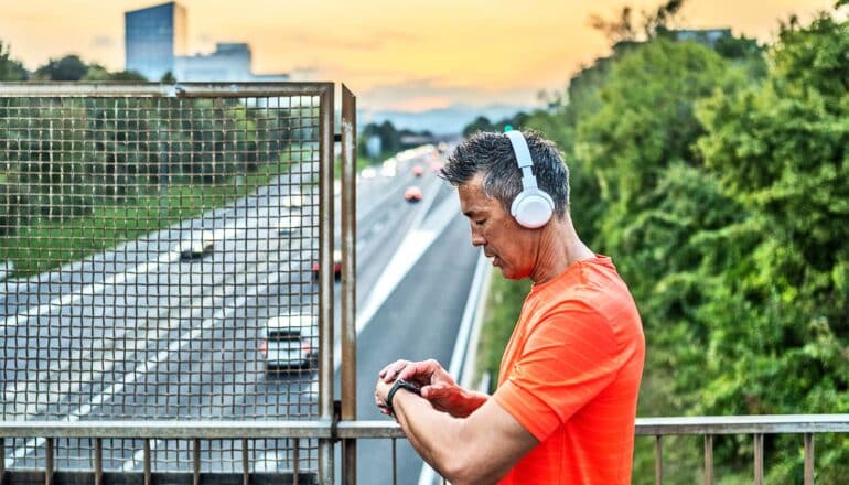 A man stops during a run to check his smartwatch while standing on a bridge over a highway lined with trees.