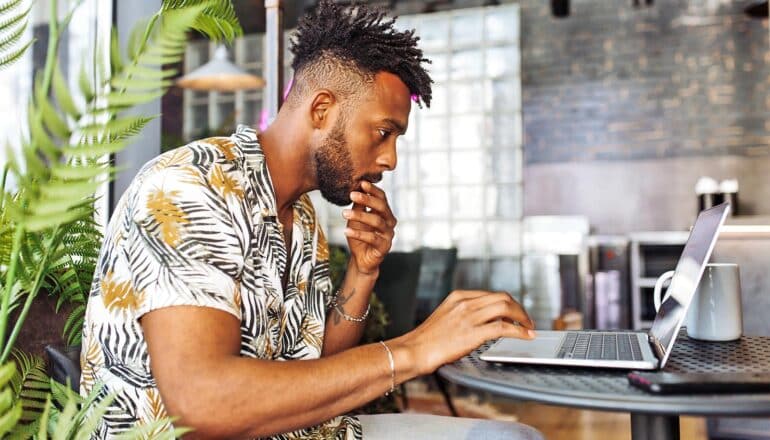 A man reads his laptop with one hand on his chin.