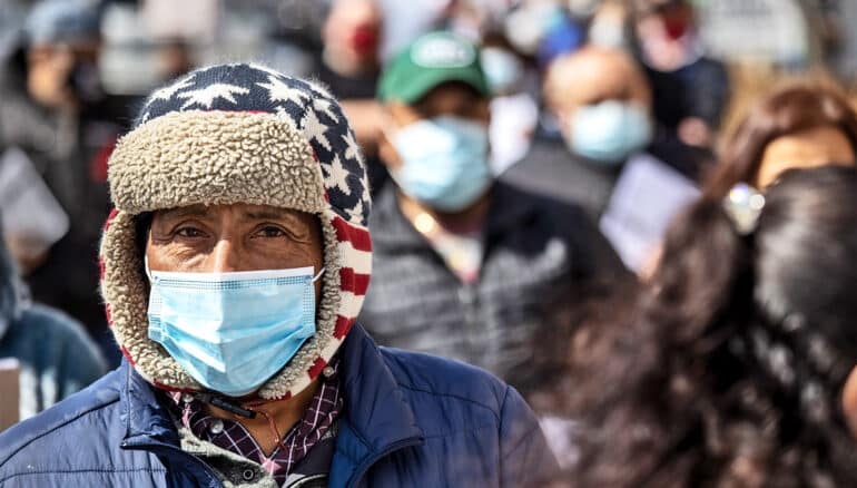 A man stands in a line wearing a mask and a winter hat with the American flag on it.