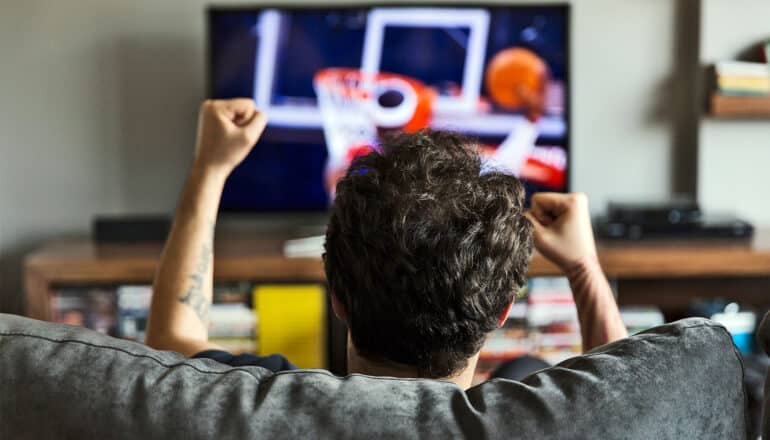 A man watches basketball on television while sitting on a couch and cheering.