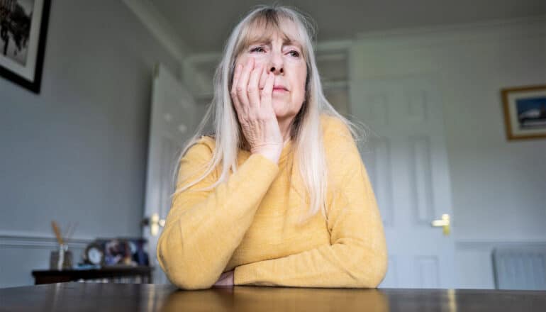 An older woman in a yellow sweater sits at a table with her hand touching her face.