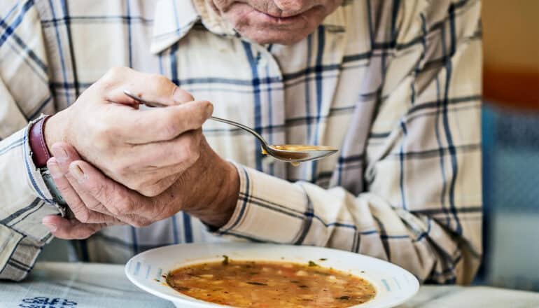 An elderly man holds a spoonful of soup in one hand and uses his other to support it.