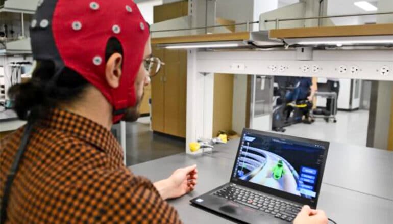 A person sits in front of a computer while wearing a cap with electrodes on it.