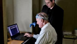 Woods stands over an older man wearing a device around his head sitting in front of a laptop.