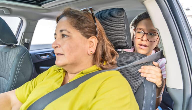 A mother drives while her daughter leans forward to talk to her from the backseat.