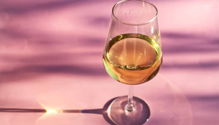 A glass of white wine sits on a pink surface.