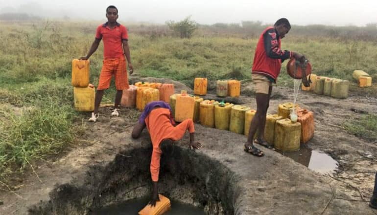 Three people gather water from a hole in large containers.