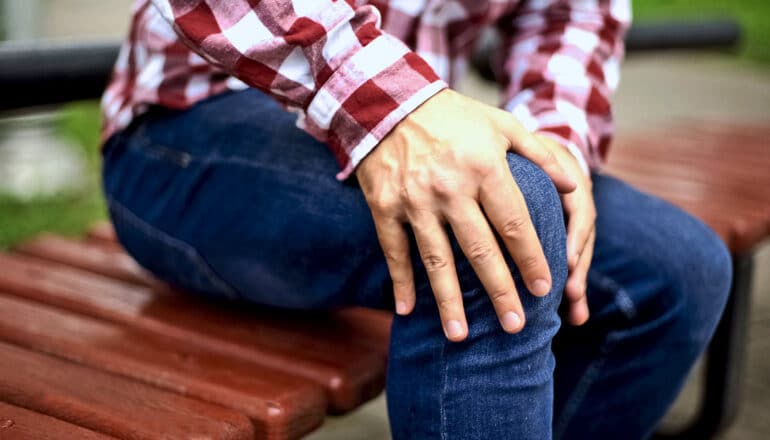 A man sitting on a park bench clutches his knee in pain.