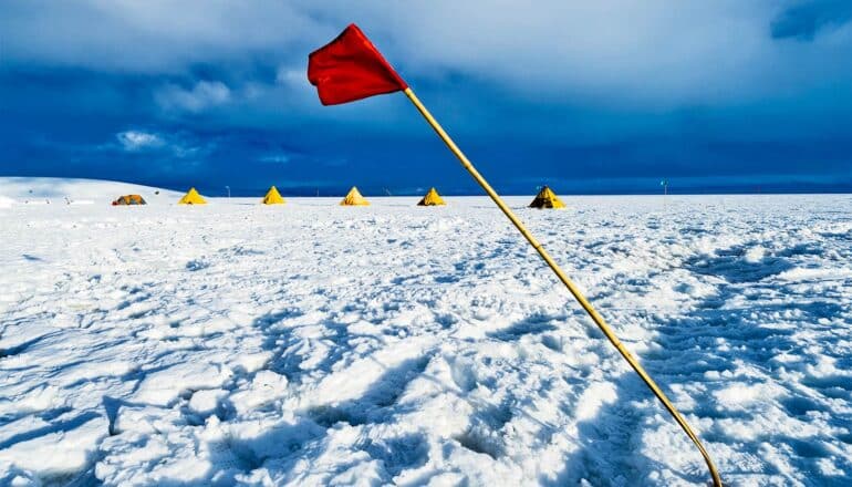 A red flag bends in the wind on the Ross Ice Shelf, with a row of yellow tents in the background.