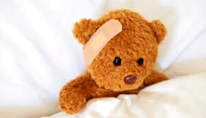 A teddy bear in bed with a bandaid on its head.