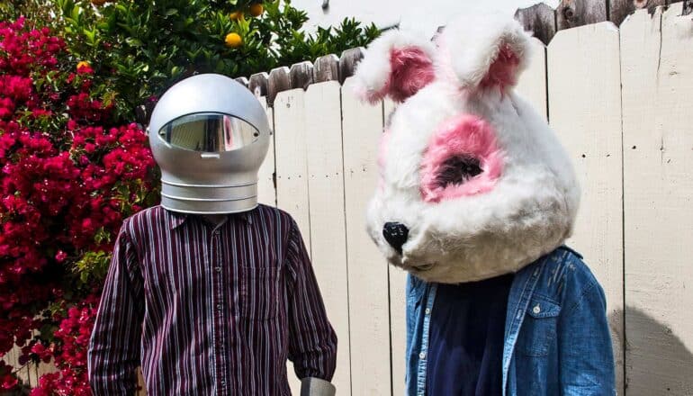 One person wears a space-age helmet and the other wears a bunny mascot head while standing in a backyard and wearing normal clothing.