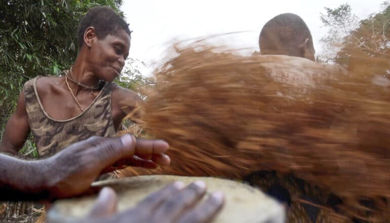 A person plays a hand drum while others dance while outside.