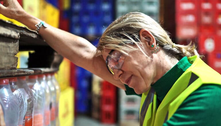 A woman working in a warehouse leans on a stack of pallets and closes her eyes.