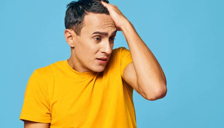 A young man in a yellow shirt looks like he's deep in thought while running his hand through his hair.