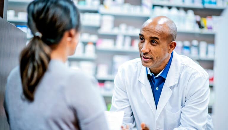 A pharmacist speaks with a customer from behind a counter.