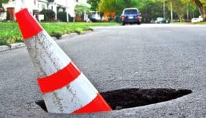 An orange safety cone sits inside a deep pothole in the road.