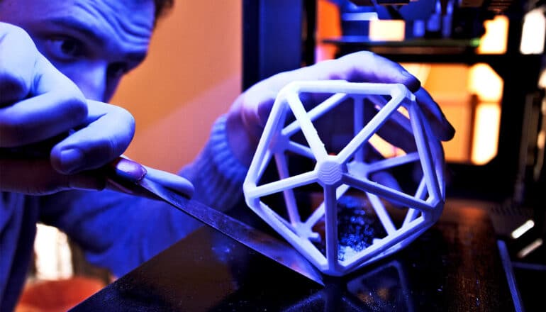 A man uses a spatula to remove a printed geometric object from a 3D printer.