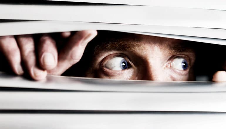 A man peeks through window blinds with a paranoid look in his eyes.