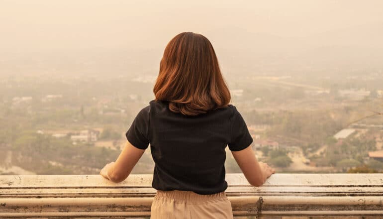 A woman with red hair looks over a balcony at a landscape covered in polluted air.