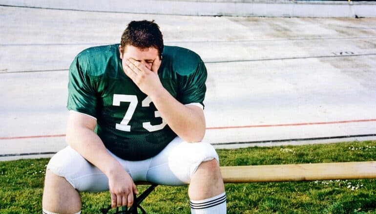 A football player touches his face while sitting on the bench.