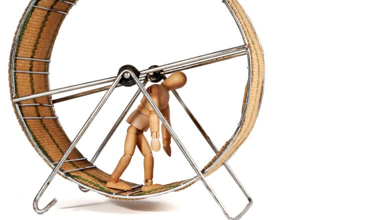 An artist's wooden model of a person walks on a hamster wheel while hunched over.