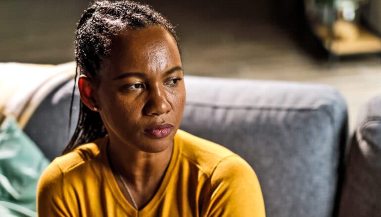 A Black woman sits on a couch while looking to her left with an anxious expression.