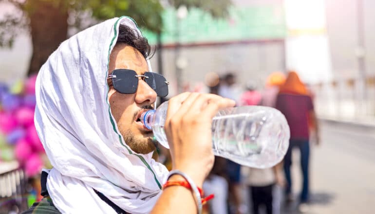 A man drinks water from a plastic bottle and wears a cloth over his head on a hot day in a city.