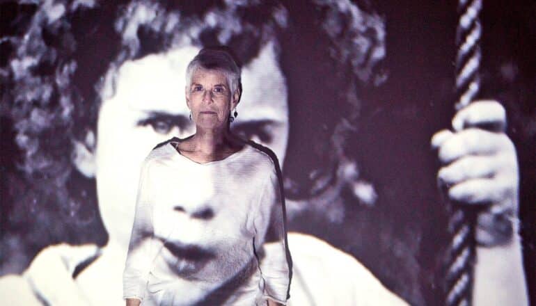 An older woman stands against a wall while a projection of a photograph showing a younger version of herself is projected onto her and the wall.