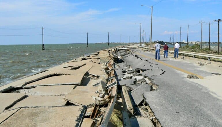 Three people walk along a heavily damaged coastal road.