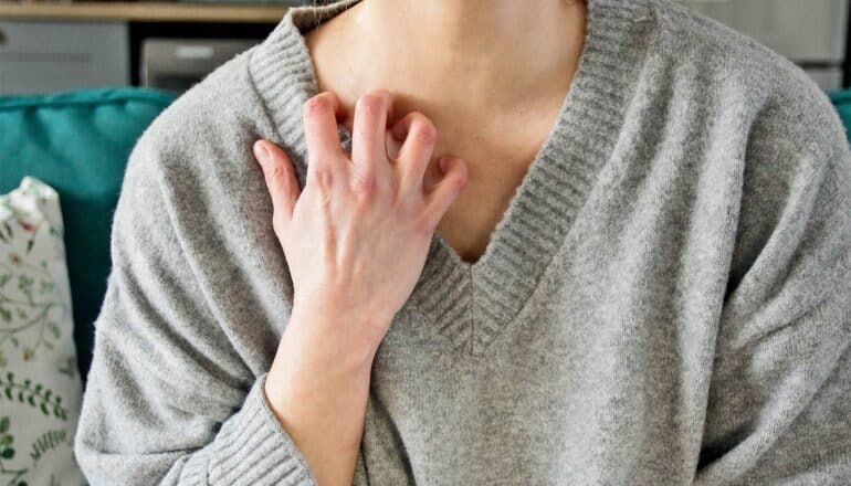 A person scratches their itchy skin on their clavicle while sitting on a couch.