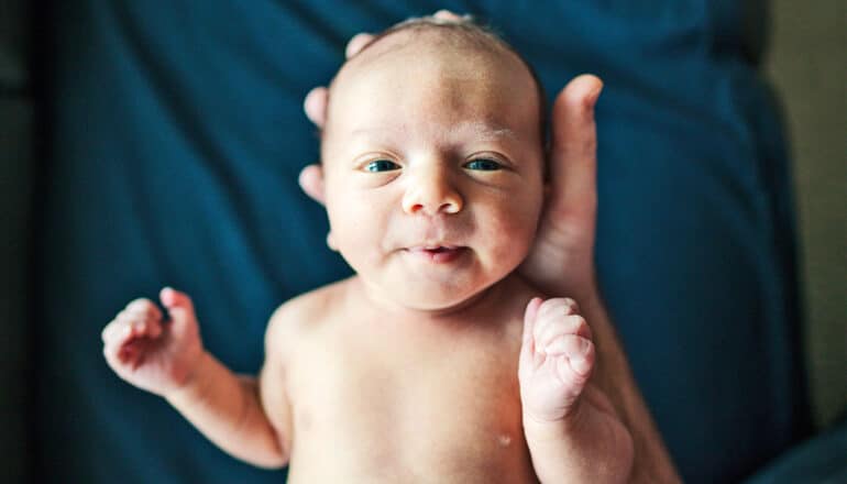 A newborn baby helds in his mother's hands looks up at the camera and smiles.