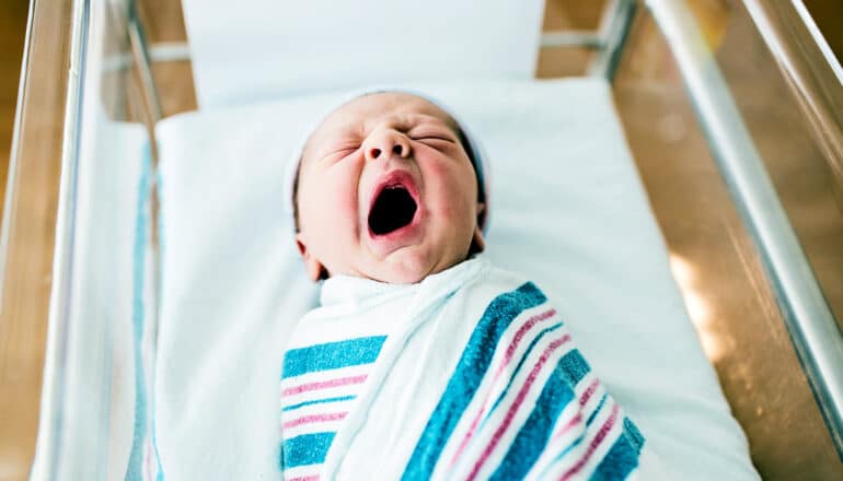 A newborn baby yawns while laying swaddled in a hospital bassinet.