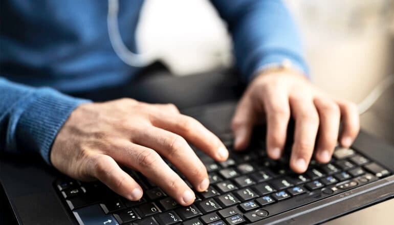 A close-up of a man's hands typing on a laptop keyboard.