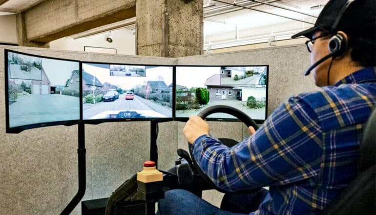 A person sits at a steering wheel in front of three computer screens showing footage capture from a car he's remotely controlling.