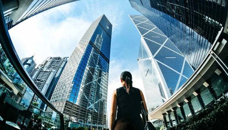 A woman walks through a city with large skyscrapers surrounding her.