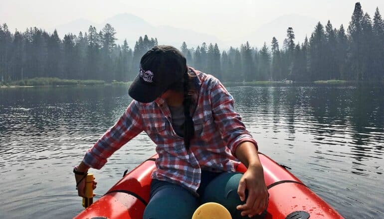 Farruggia dips an instrument in the lake while sitting on a red dingy as smoke obscures mountains around the lake.